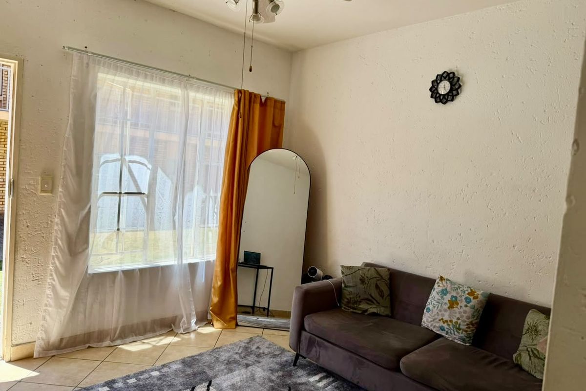 A living room featuring tiled flooring, a ceiling fan, and a large window with security gate, offering a bright and functional living space.