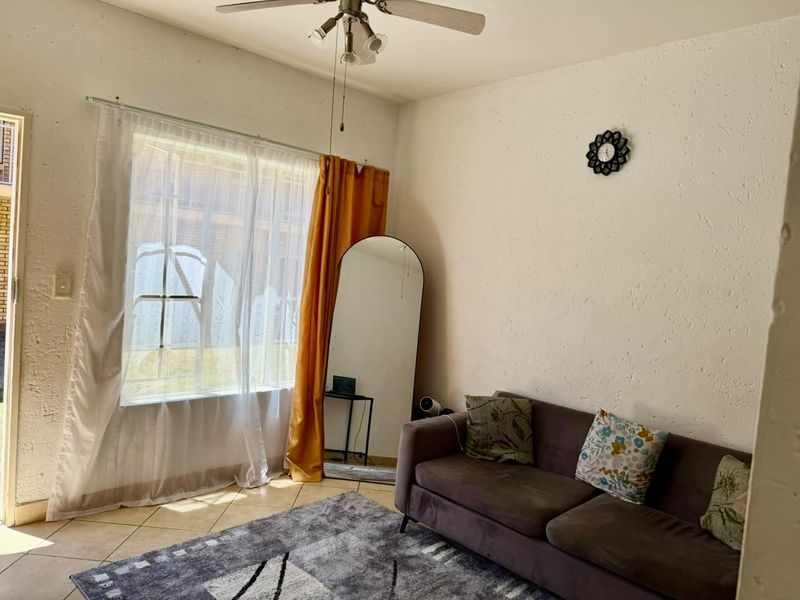 A living room featuring tiled flooring, a ceiling fan, and a large window with security gate, offering a bright and functional living space.