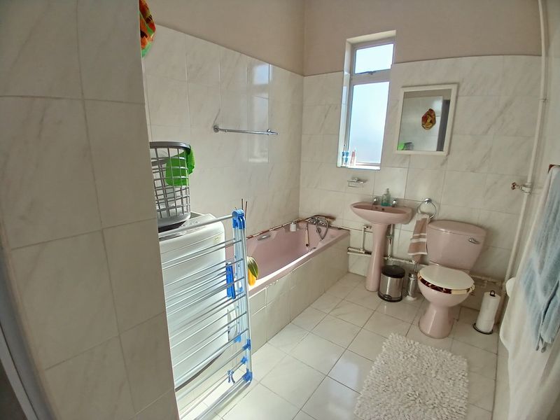 A dated bathroom featuring a pink bathtub, pedestal basin, and toilet with full-height wall tiling and tiled flooring.