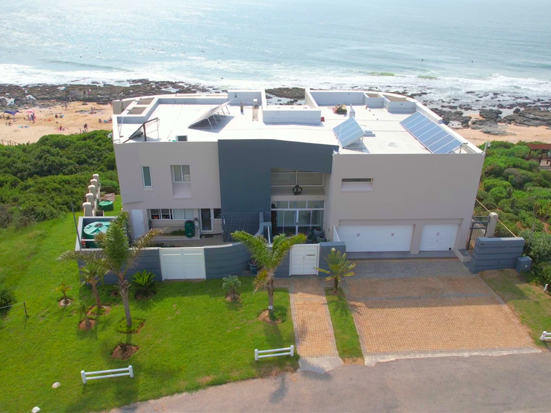 Aerial view of a modern beachfront property featuring a flat roof with solar panels, a double garage, and a paved driveway.