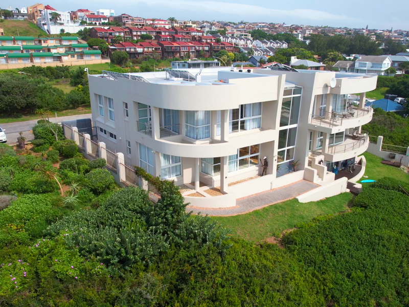 Aerial view of a modern multi-level residential property featuring curved balconies, large glass windows, and solar panels, set within a landscaped coastal environment.
