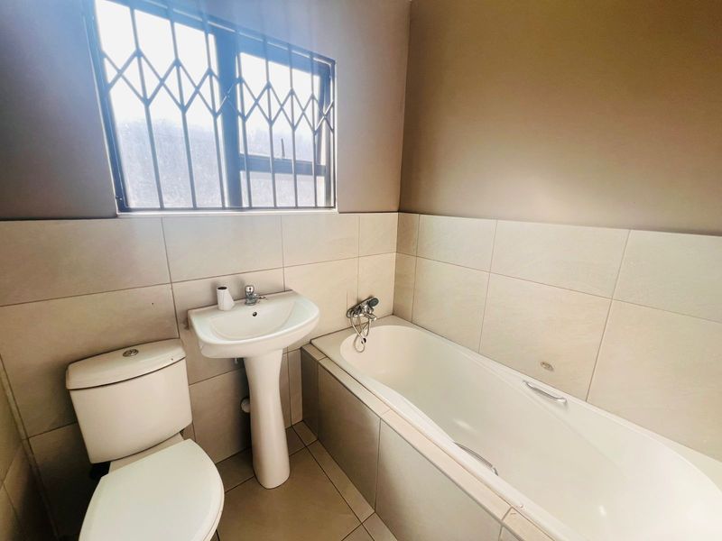 A functional bathroom featuring a pedestal basin, toilet, and bathtub with tiled finishes and a window with security bars.