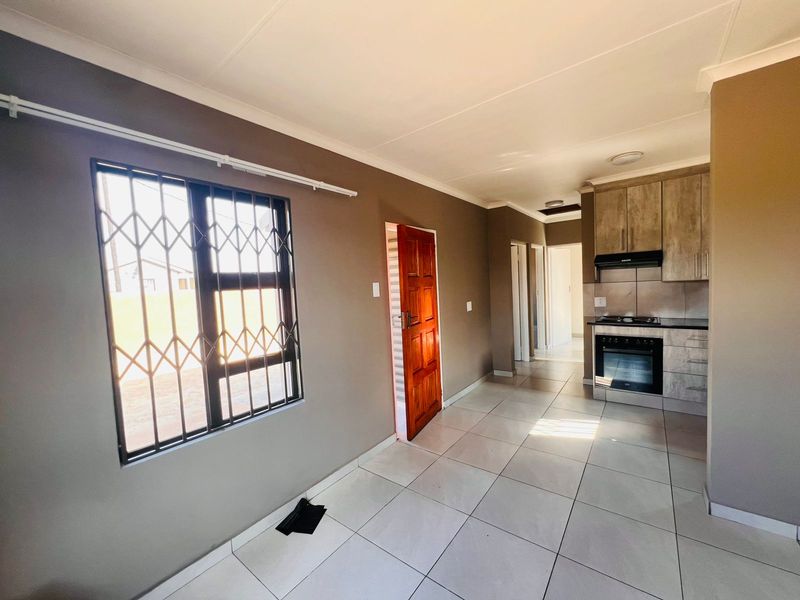 Open plan living and kitchen area featuring tiled flooring, built-in cabinetry with integrated oven and hob, and a secure window with burglar bars.