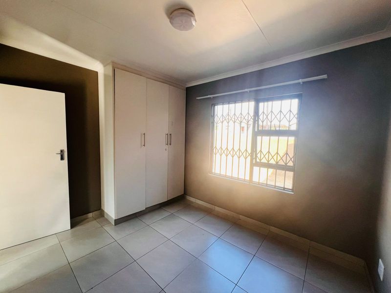 A clean, empty bedroom featuring tiled flooring, built-in cupboards, and a window fitted with a security gate.