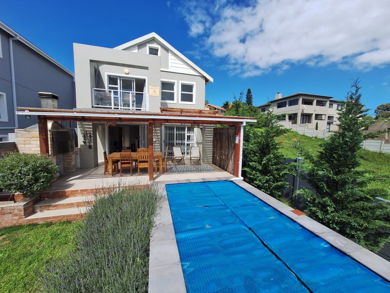 Rear exterior view featuring a covered patio with built-in braai, a swimming pool with cover, and an upper-level balcony.