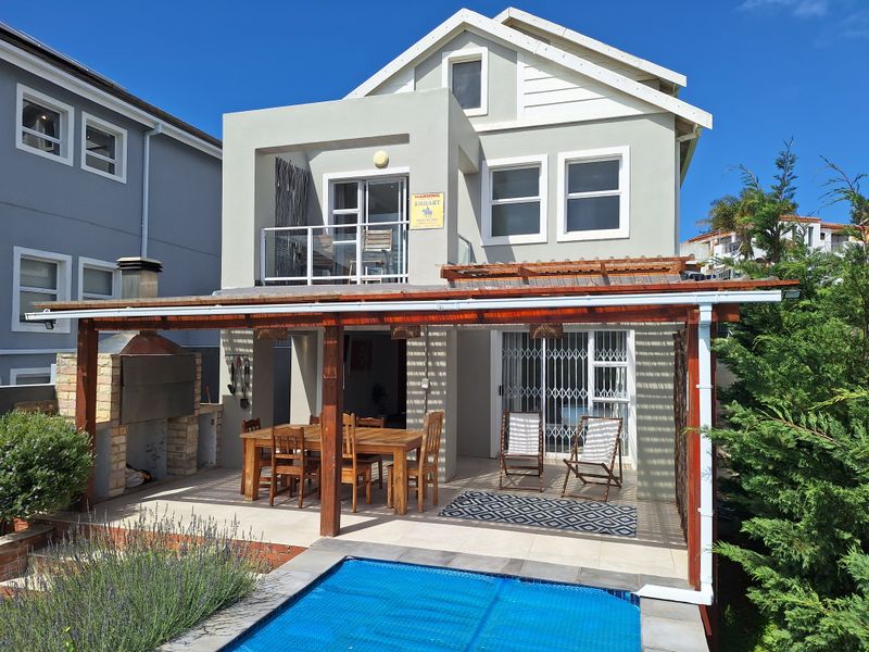 Rear exterior view featuring a covered patio with a built-in braai, swimming pool, and an upper-level balcony.