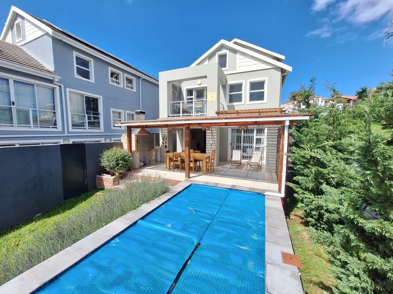 Rear exterior view featuring a swimming pool with cover, a covered patio area with a built-in braai, and an upper-level balcony.