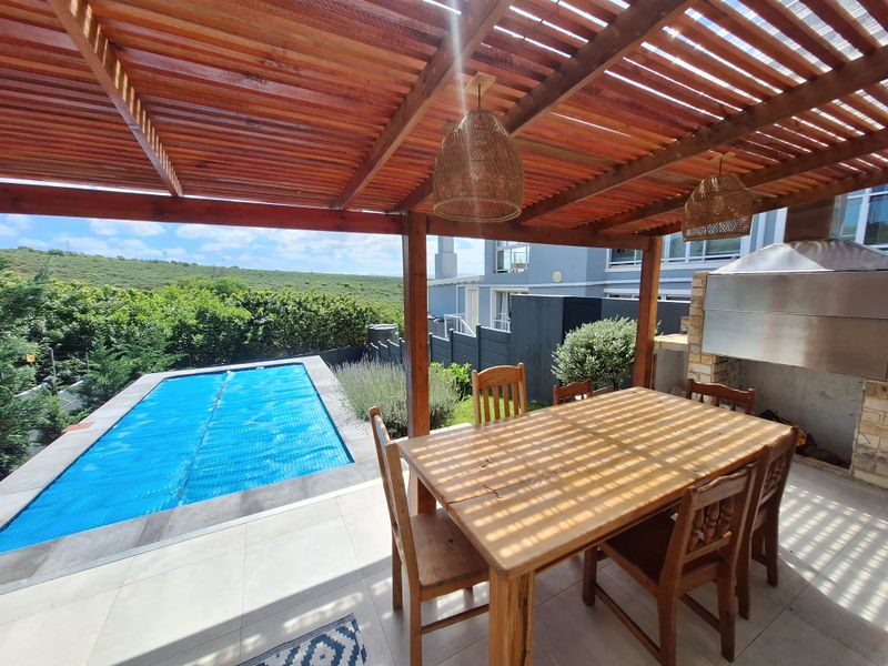 Covered outdoor patio featuring a wooden pergola, built-in braai, and tiled flooring, overlooking a private swimming pool and landscaped garden area.