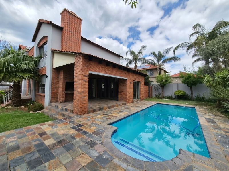 A well-maintained outdoor entertainment area featuring a swimming pool, stone-paved patio, and a brick-built braai structure attached to the main house.