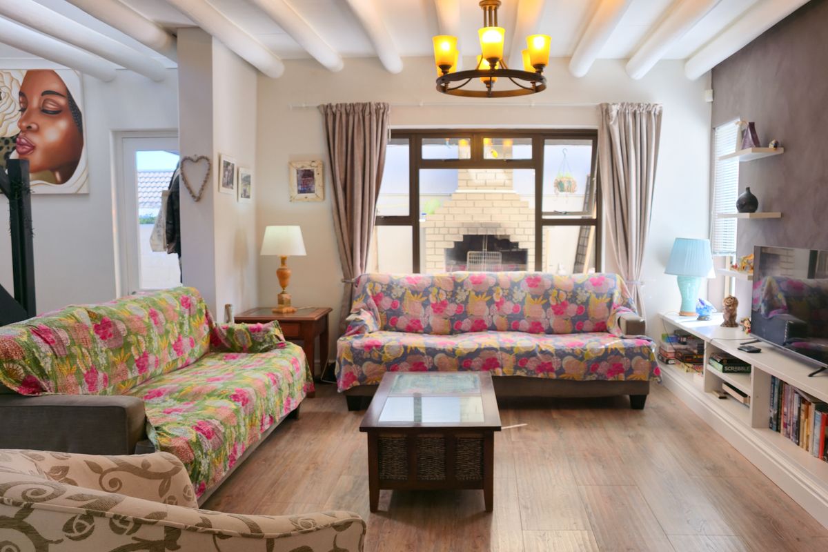 A cozy living room featuring exposed ceiling beams, laminate flooring, and built-in shelving, with a view through the window to an outdoor brick fireplace.