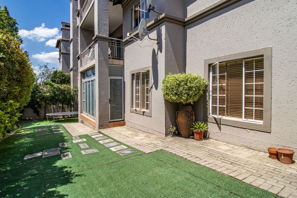 Rear exterior view of the apartment featuring a paved patio area, artificial lawn, and access to a balcony with metal railings.