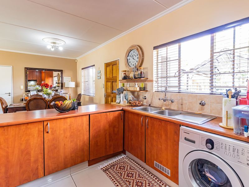 Open-plan kitchen area featuring wooden cabinetry, a double stainless steel sink, tiled backsplash, and integrated laundry plumbing, flowing into the adjacent dining space.