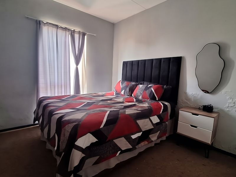A bedroom featuring a window with a curtain rod, showing significant wall damage and worn carpeting requiring maintenance.
