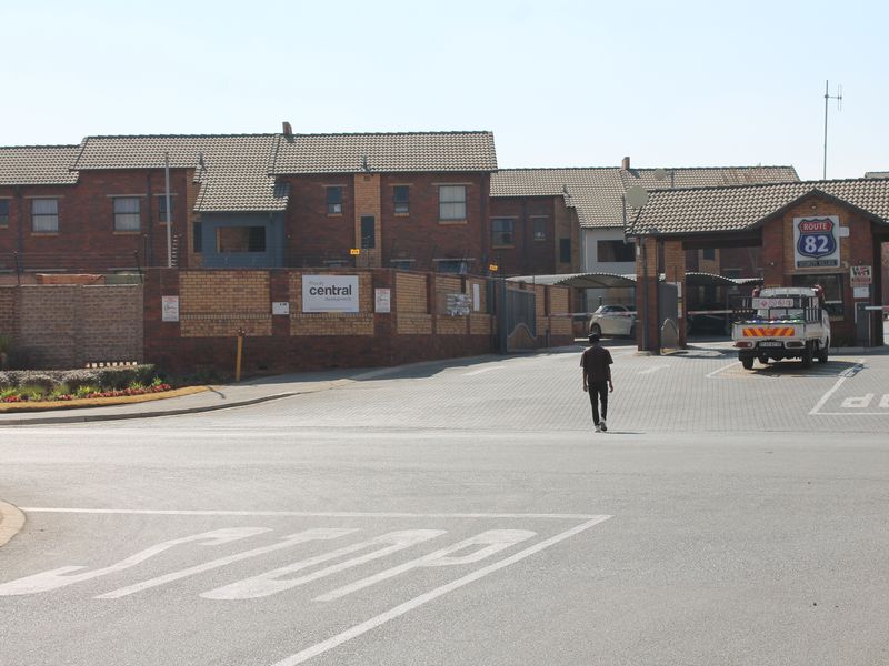 Street view of the entrance to the Route 82 security village complex, featuring brick facade buildings, paved access roads, and a secure gatehouse.