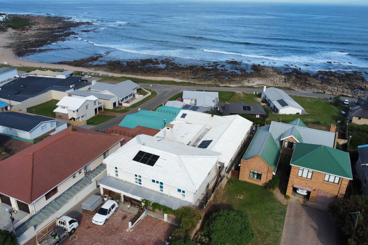Aerial view of a coastal property featuring a white metal roof with solar panels, situated near the ocean with a paved driveway.