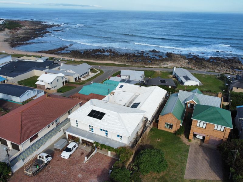 Aerial view of a coastal property featuring a white metal roof with solar panels, situated near the ocean with a paved driveway.