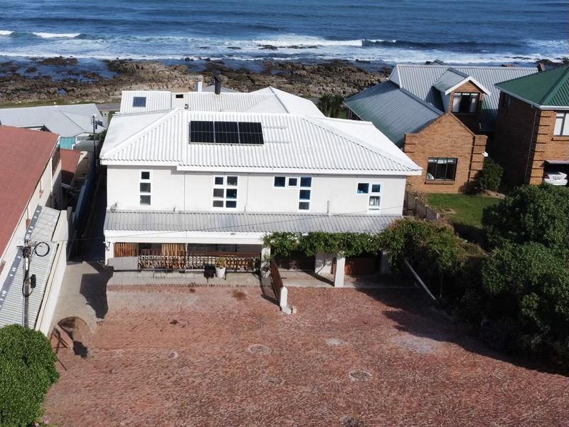 Aerial view of a coastal property featuring a white metal roof with solar panels, a spacious paved driveway, and a covered patio area.