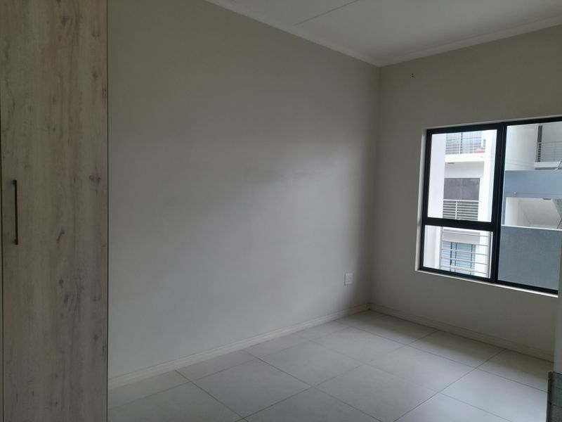 A bright, empty bedroom featuring neutral tiled flooring, a modern built-in wardrobe, and a large window with an aluminium frame.