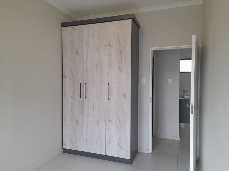 A clean, empty bedroom featuring a modern built-in wardrobe with wood-grain finish and neutral tiled flooring, leading to an adjacent room.