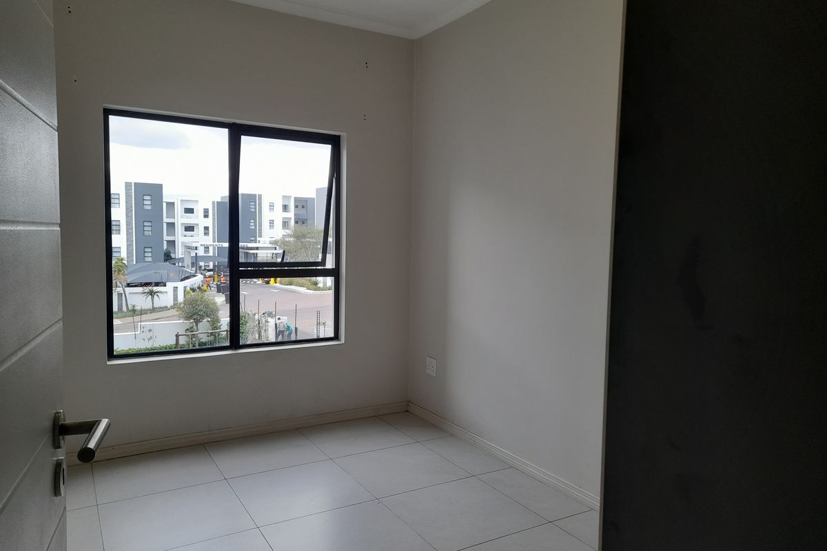 A bright, empty bedroom featuring neutral tiled flooring and a large aluminium-framed window with views of the surrounding apartment complex.
