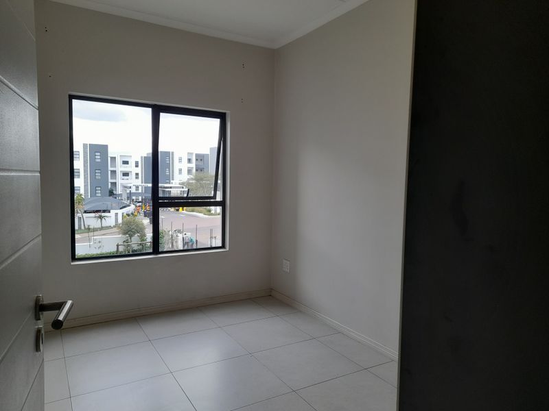 A bright, empty bedroom featuring neutral tiled flooring and a large aluminium-framed window with views of the surrounding apartment complex.