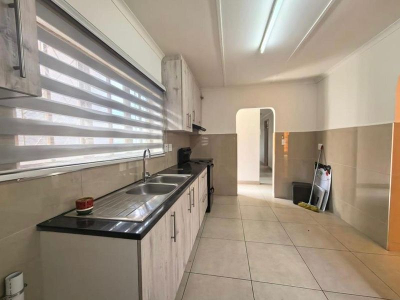 A functional kitchen featuring light wood-look cabinetry, a double basin sink, tiled splashbacks, and neutral floor tiles with an open archway leading to adjacent areas.