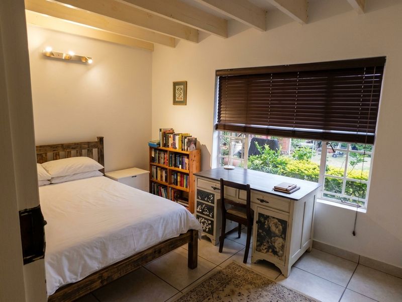 A cozy bedroom featuring exposed timber ceiling beams, tiled flooring, and a large window with venetian blinds overlooking the garden.