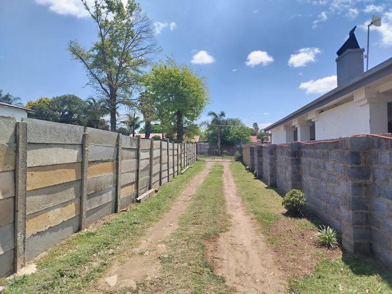A rear exterior view showing a dirt driveway flanked by precast concrete and cinder block boundary walls leading toward the back of the property.