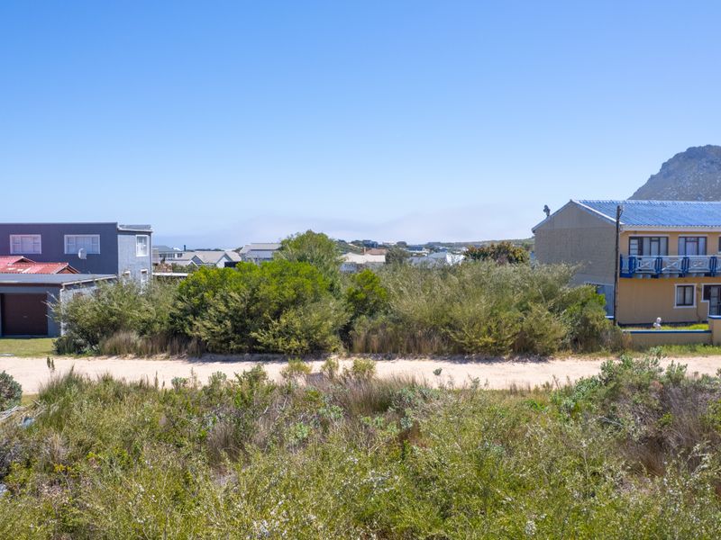 A view of a vacant residential plot featuring natural coastal vegetation and sandy terrain, situated in a developed neighborhood with surrounding homes.