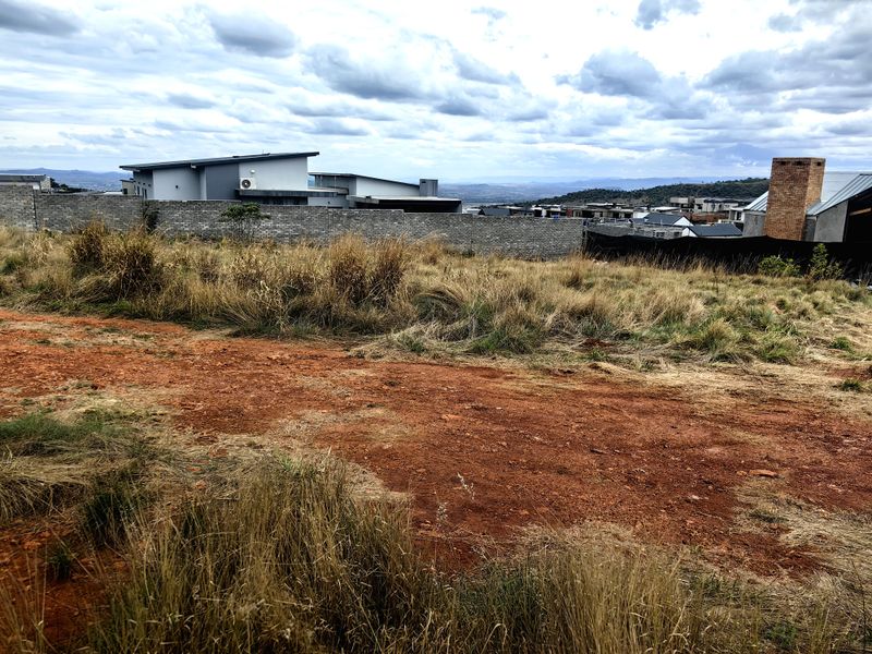A vacant residential plot featuring natural grass cover, an unpaved access path, and a perimeter boundary wall in a developed estate setting.