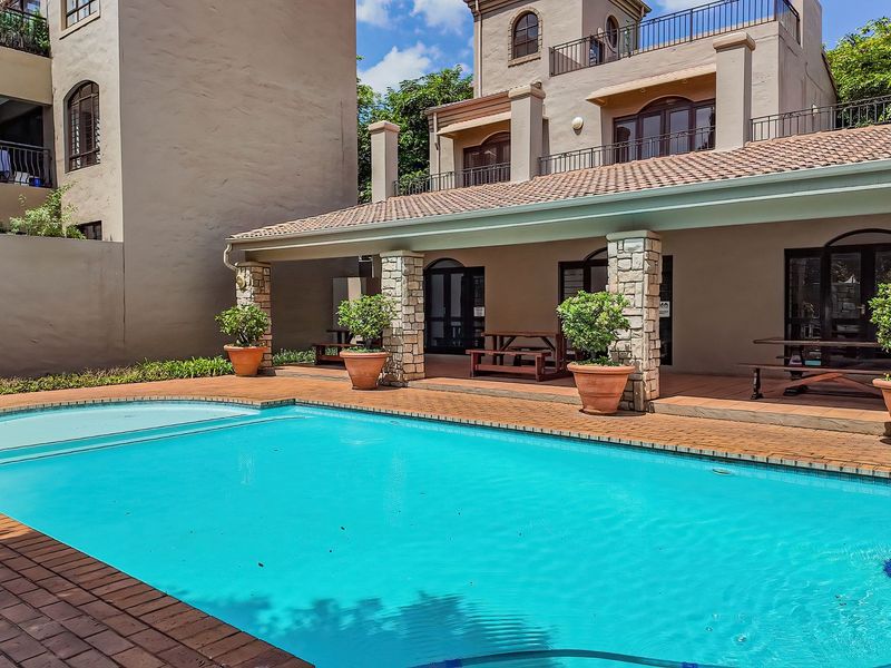 A well-maintained outdoor pool area featuring a swimming pool, brick paving, and a covered patio with stone-clad pillars.