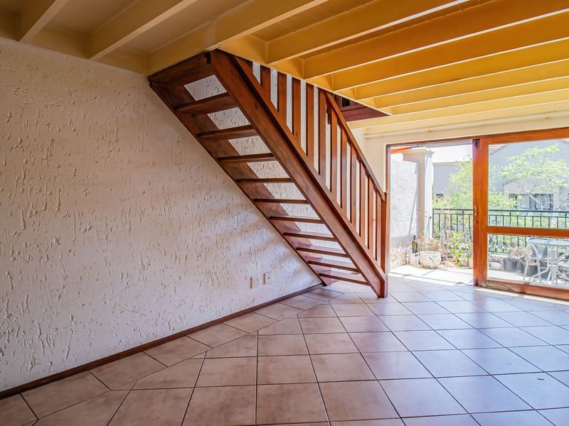 An open-plan living area featuring a wooden staircase, exposed ceiling beams, and tiled flooring, with direct access to an outdoor patio area.