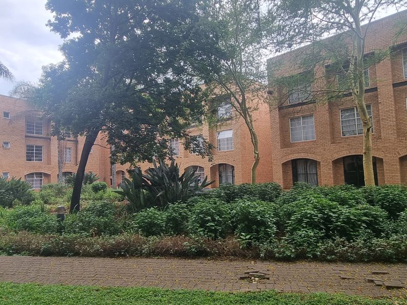 A view of the rear exterior of a multi-story face brick apartment complex featuring a landscaped garden and paved walkway.