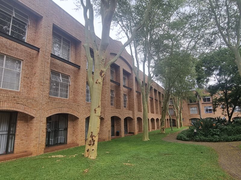 Exterior view of a multi-story apartment complex featuring a face brick facade, arched ground floor openings, and a well-maintained garden with mature trees.