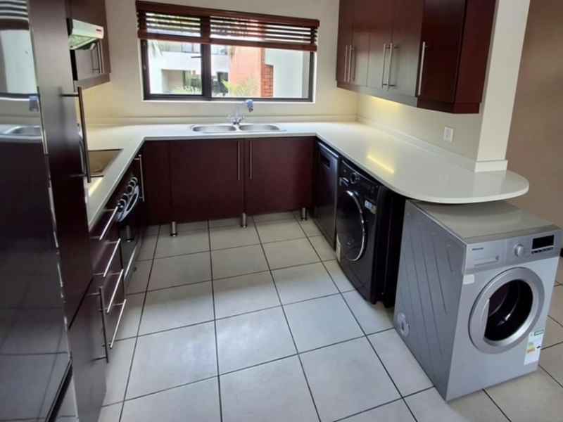 A functional kitchen featuring dark wood cabinetry, light countertops, tiled flooring, and a window with venetian blinds, offering a clean and practical workspace.