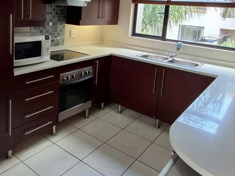 A functional kitchen featuring dark wood cabinetry, white stone countertops, a stainless steel double sink, and built-in appliances with a tiled backsplash.