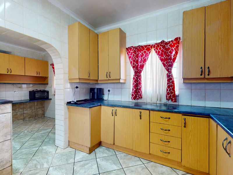 A functional kitchen featuring light wood-look cabinetry, dark laminate countertops, tiled backsplash, and tiled flooring, connected to an adjacent prep area via an archway.