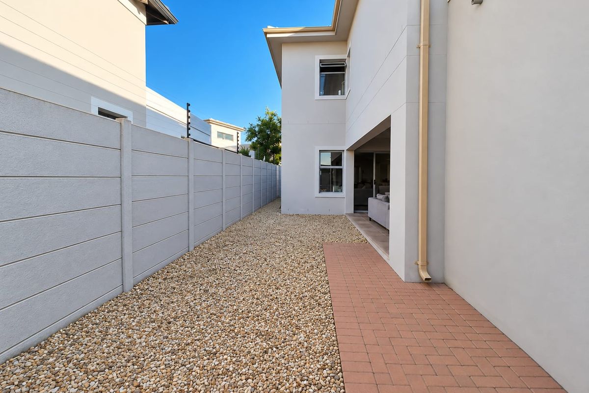 A clean and well-maintained rear exterior area featuring a paved walkway, pebble landscaping, and a secure boundary wall with electric fencing.
