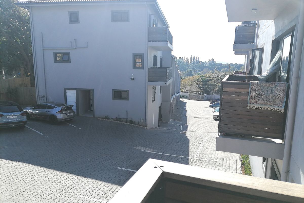 View of the apartment complex exterior, featuring a paved driveway, parking area, and private balconies with timber railings.
