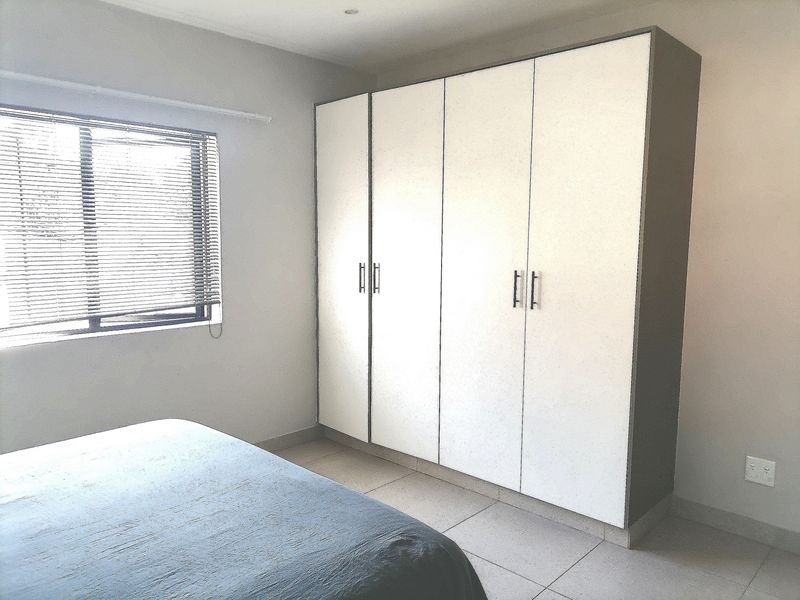 A bright bedroom featuring neutral tiled flooring, a large built-in wardrobe with white doors, and venetian blinds on the window.