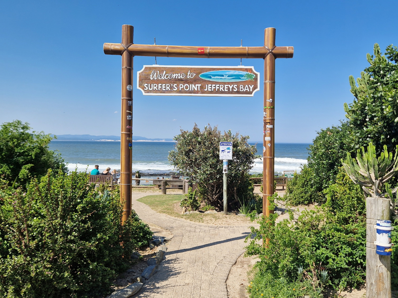 A scenic view of the ocean at Surfer's Point in Jeffreys Bay, featuring a welcoming entrance sign and a paved pathway leading to the beach.