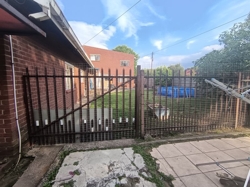 Rear exterior view of the property featuring a brick facade, metal security fencing, and a paved patio area leading to a lawn.