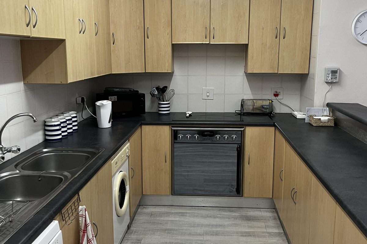 A functional kitchen featuring light wood-finish cabinetry, dark laminate countertops, a built-in oven with hob, and tiled splashbacks with integrated appliance connections.