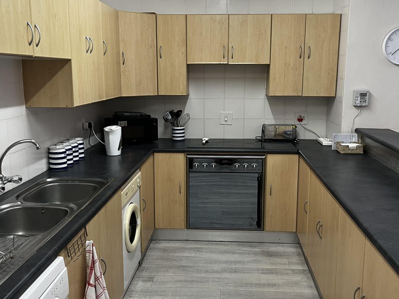 A functional kitchen featuring light wood-finish cabinetry, dark laminate countertops, a built-in oven with hob, and tiled splashbacks with integrated appliance connections.
