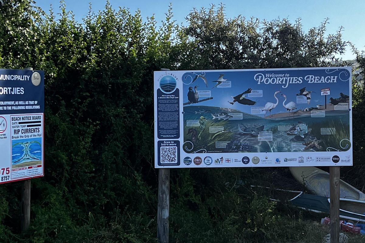 An outdoor area featuring informational signage for Poortjies Beach, set against natural coastal vegetation and sandy terrain.