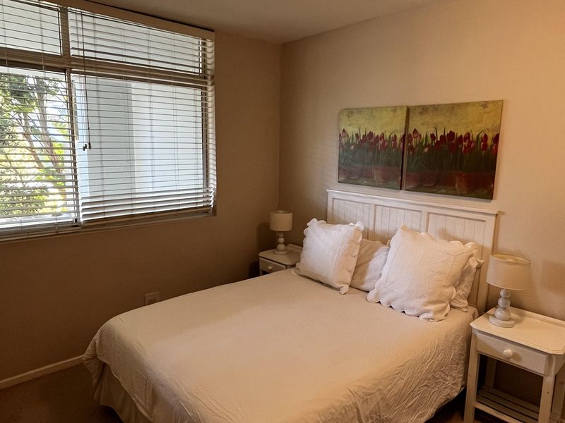A cozy bedroom featuring neutral wall tones, carpeted flooring, and large windows fitted with venetian blinds, providing natural light.