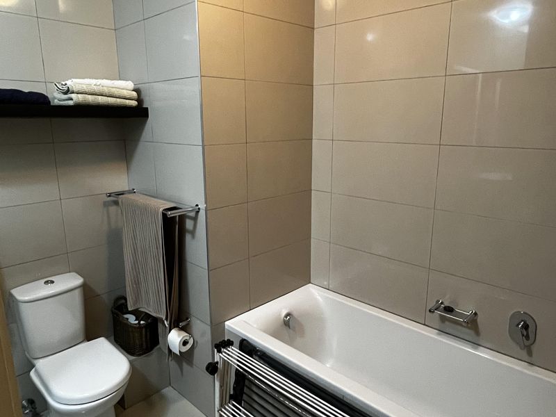 A functional bathroom featuring a built-in bathtub, toilet, and neutral ceramic tiling throughout, providing a clean and practical space.