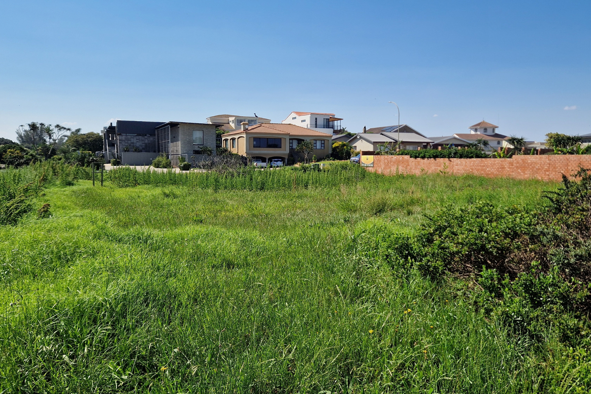 A vacant residential plot covered in grass, featuring a brick boundary wall and surrounding neighborhood homes in the background.