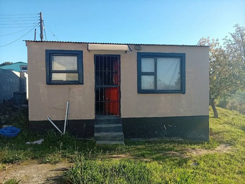 A modest exterior view of a small dwelling featuring a corrugated roof, security gate, and concrete entrance steps, requiring exterior maintenance and painting.