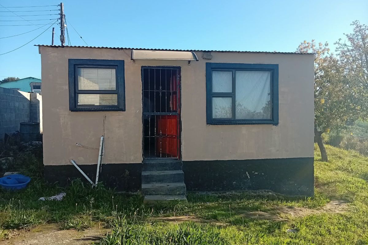 A modest exterior view of a small dwelling featuring a corrugated roof, security gate, and concrete entrance steps, requiring exterior maintenance and painting.